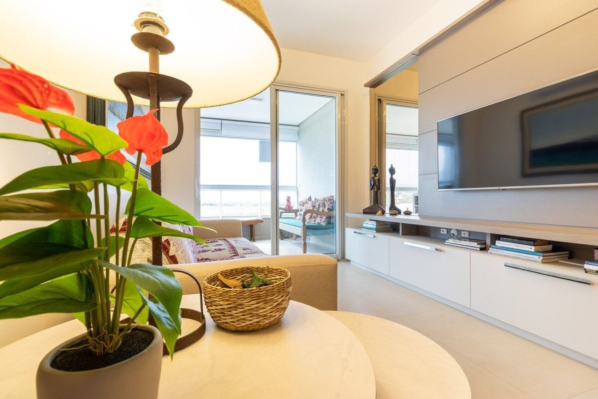 A cozy living area features a soft sofa and a round table adorned with a decorative lamp and a vibrant plant. A television is mounted on the wall opposite a large window, which opens to a balcony with seating.