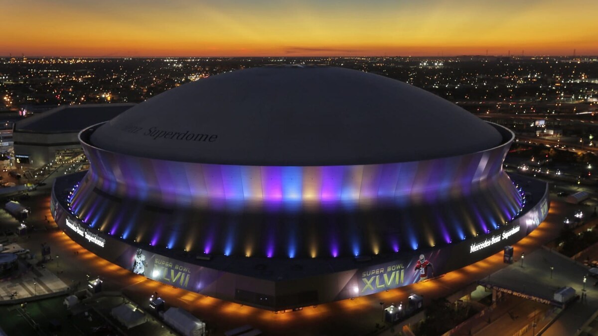 The image captures the Superdome's iconic structure illuminated with vibrant colors against a twilight sky. The surrounding cityscape is visible, with lights twinkling in the distance as the sun sets, creating a serene ambiance.