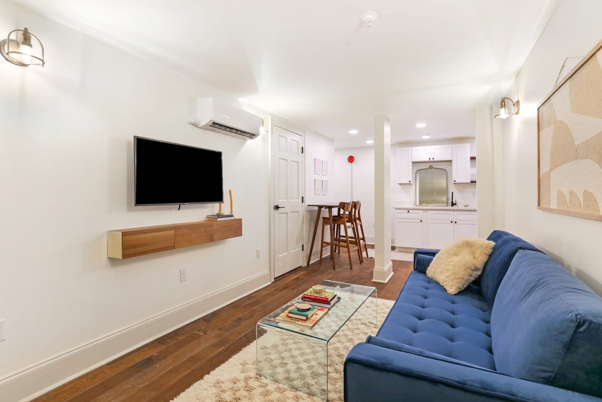 A modern living area is presented, featuring a blue sofa and a glass coffee table. A wall-mounted television is positioned above a wooden console. The space connects to a kitchen area, which is visible in the background, and soft ambient lighting enhances the inviting environment.