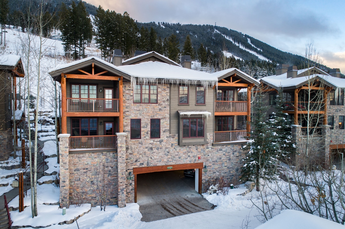 A multi-level stone building is situated in a snowy landscape. Wood accents frame the windows and balconies, while the mountain backdrop adds a natural element to the setting. An underground garage entrance is visible at the front of the property.