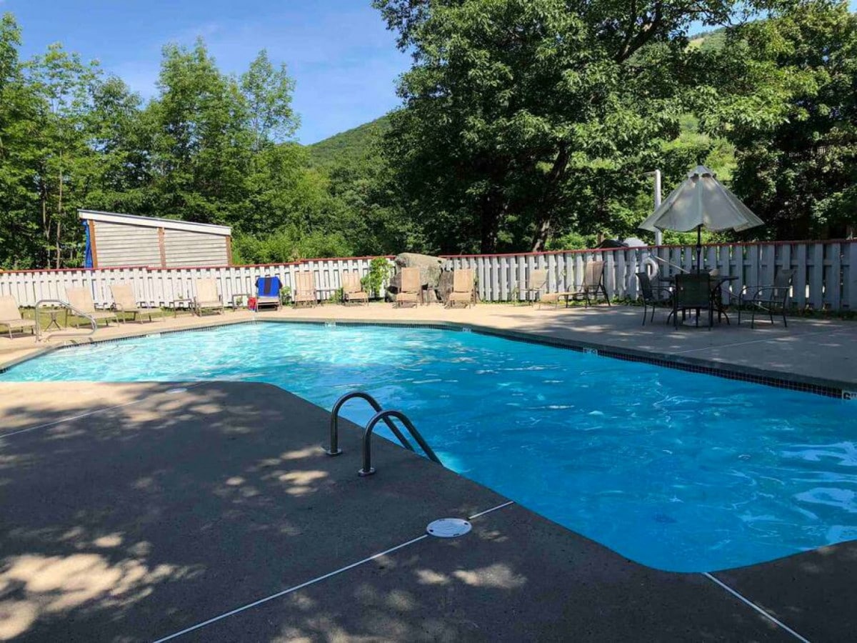 A clear blue pool is surrounded by several lounge chairs and tables under the shade of trees. The area is enclosed with a white fence, and a patio umbrella offers additional shade. Lush greenery can be seen in the background.
