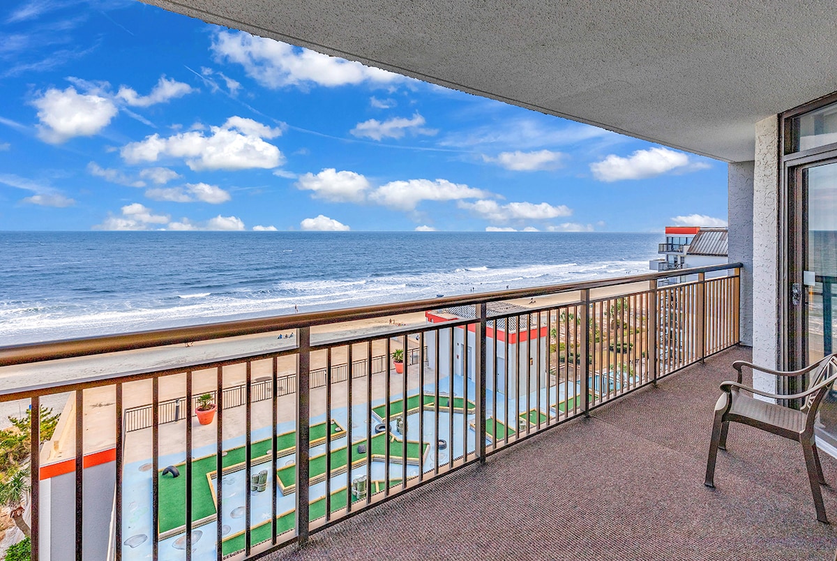 A spacious balcony provides a panoramic view of the ocean, showcasing gentle waves and a clear blue sky. The railing complements the scenery, while outdoor seating is available for relaxation. Below, the resort's pools can be seen, enhancing the coastal atmosphere.