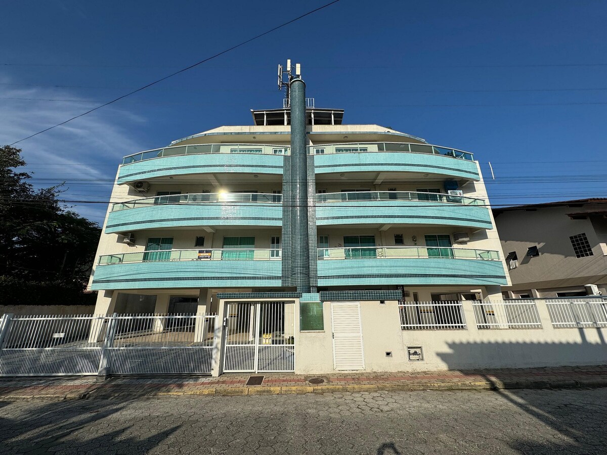 A multi-story building with a modern design features a light-colored façade and multiple balconies surrounded by a turquoise railing. A central column runs vertically along the front, while a secure entry gate is visible at the base. A clear blue sky serves as the backdrop.