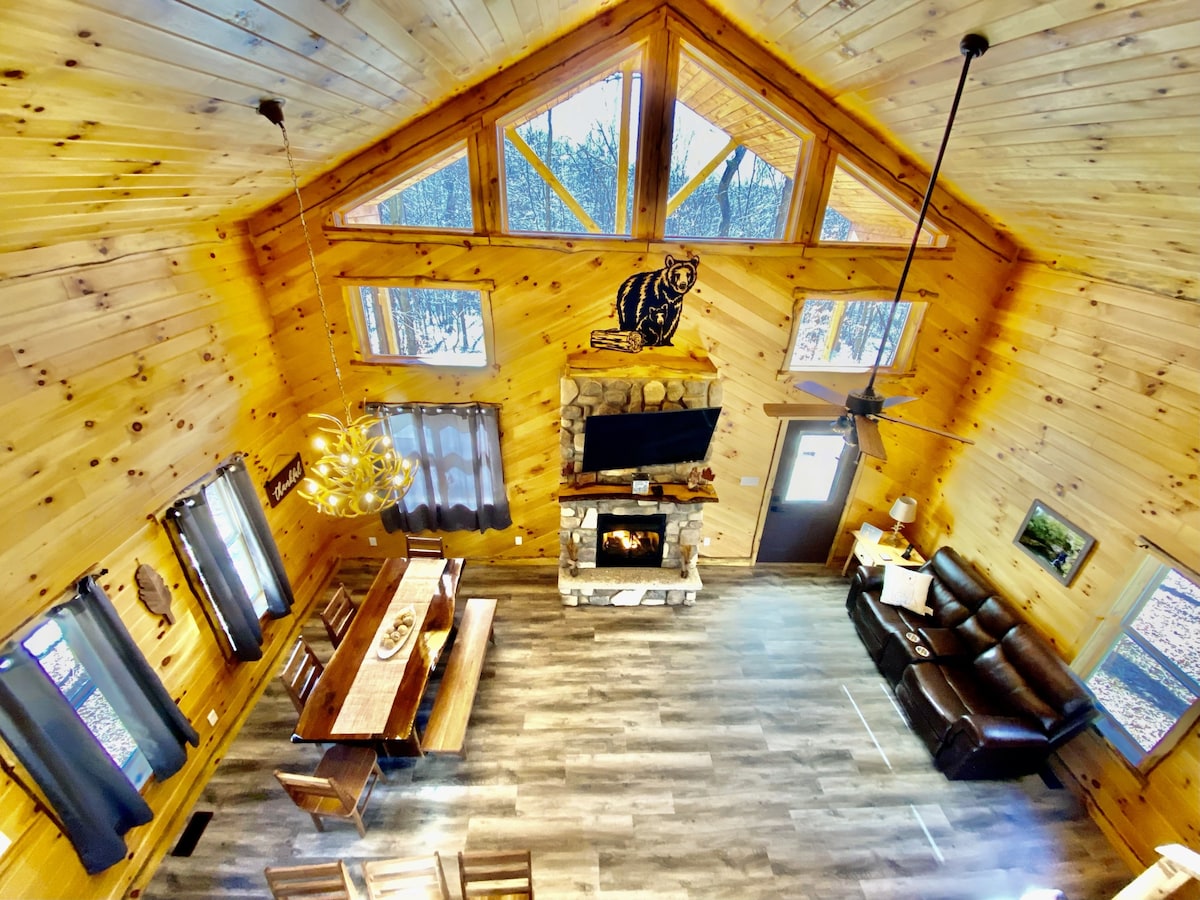 An aerial view of the spacious living area within the log cabin showcases warm wooden walls and a high vaulted ceiling. Natural light floods in through large windows. A stone fireplace is centered at the back, complemented by a leather seating area and a long dining table. Decorative accents, including a bear figure, enhance the rustic charm.