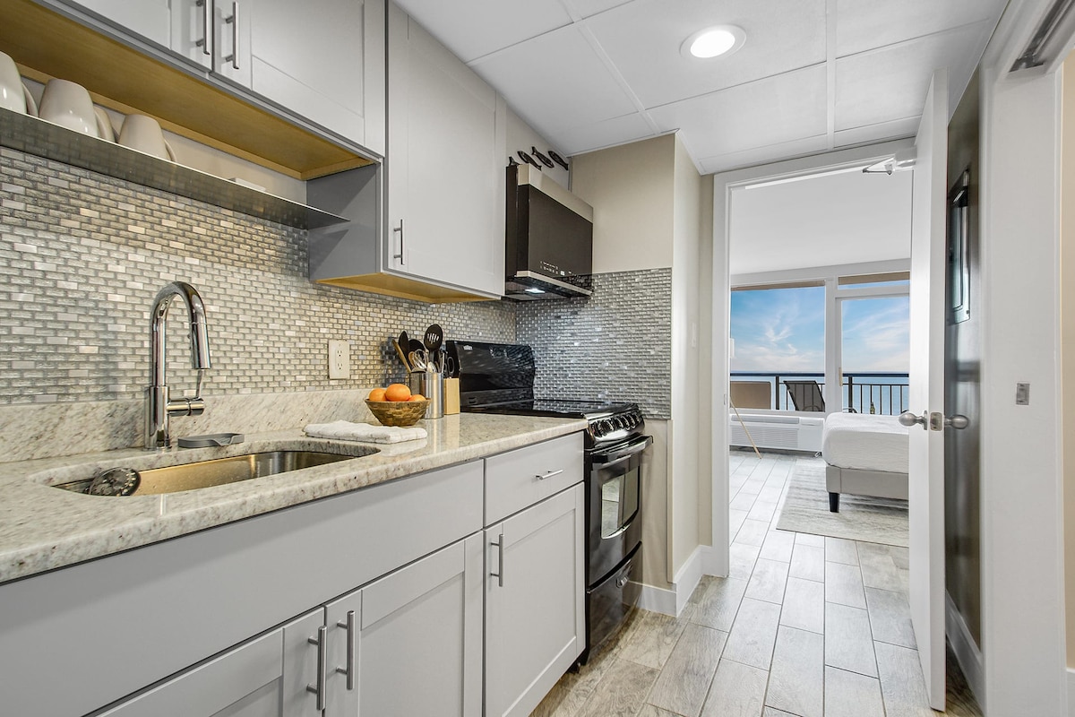 A modern kitchen features sleek gray cabinetry and light gray quartz countertops. An attractive backsplash complements the design, while essential appliances, including a stove and microwave, are visible. The kitchen allows for easy access to the open living area and private balcony with ocean views.
