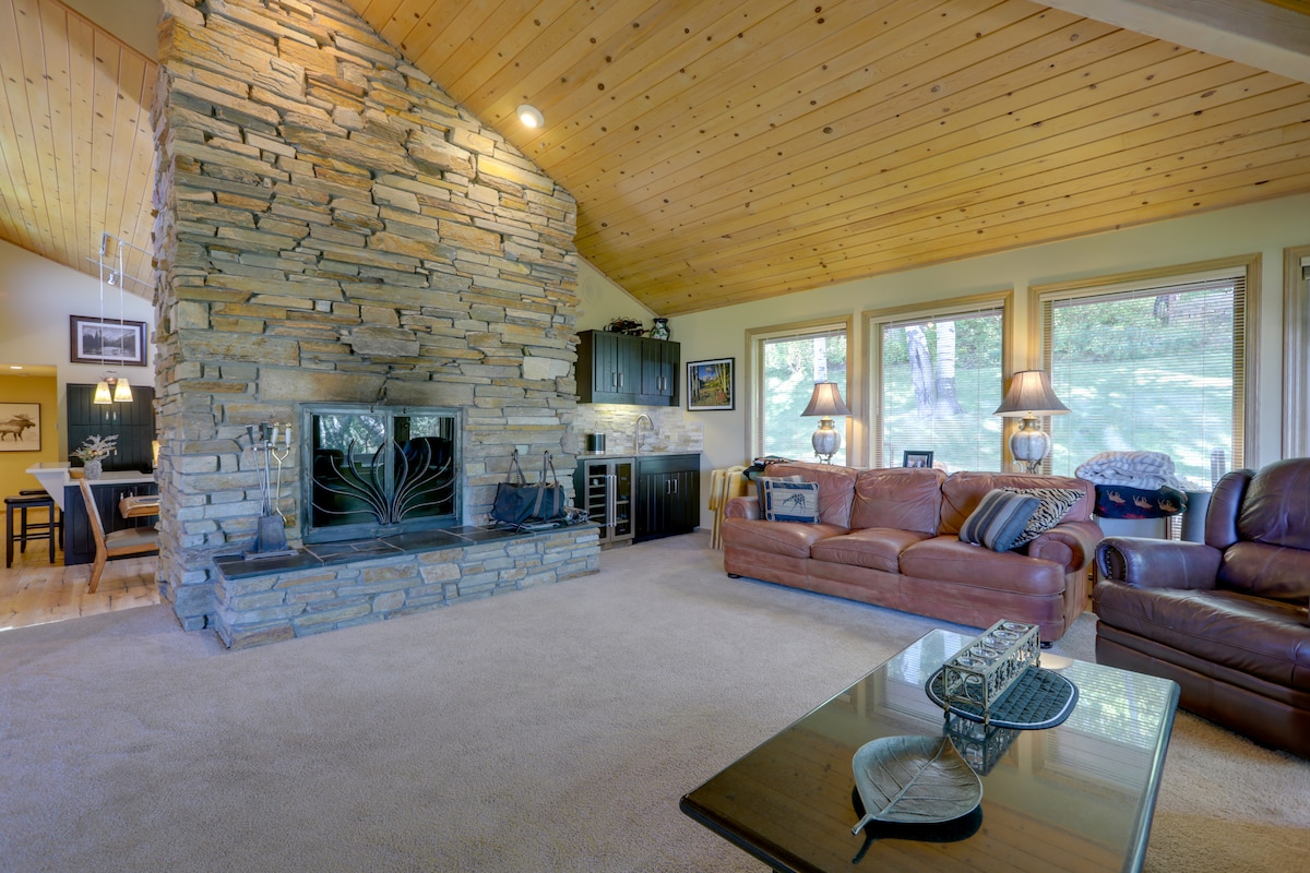The spacious living area features a large stone fireplace and wood paneling on the ceiling. Comfortable leather sofas surround a glass coffee table, while large windows provide views of the outdoors. A dining area is visible in the background.