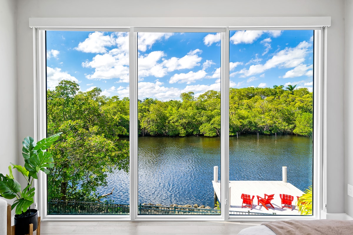 A large window reveals views of a serene river framed by lush greenery. A wooden deck extends over the water, with red outdoor chairs positioned for relaxation. The sky is dotted with fluffy clouds, enhancing the natural light that fills the space.