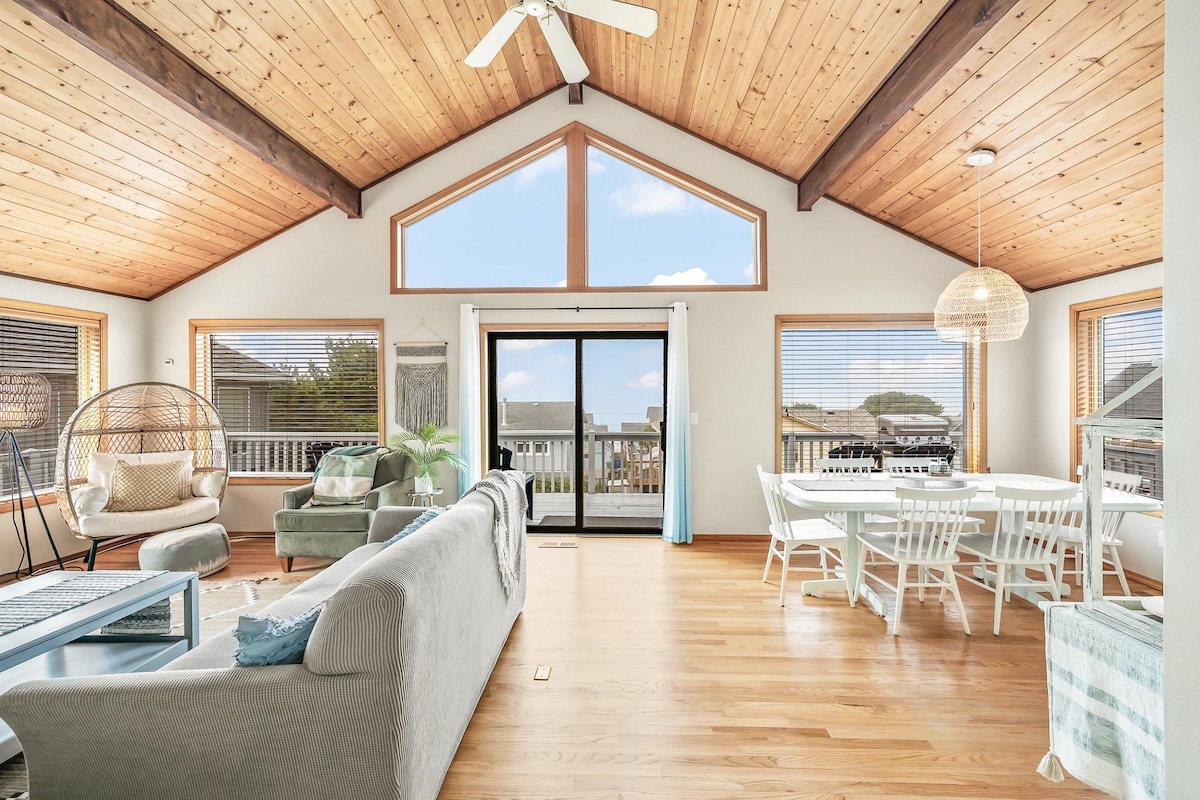 A spacious living area showcases a wood plank ceiling and large windows allowing ample natural light. Comfortable couches and a stylish dining table provide seating for guests. Sliding doors open to a balcony, and a unique hanging chair adds a distinctive touch to the coastal ambiance.