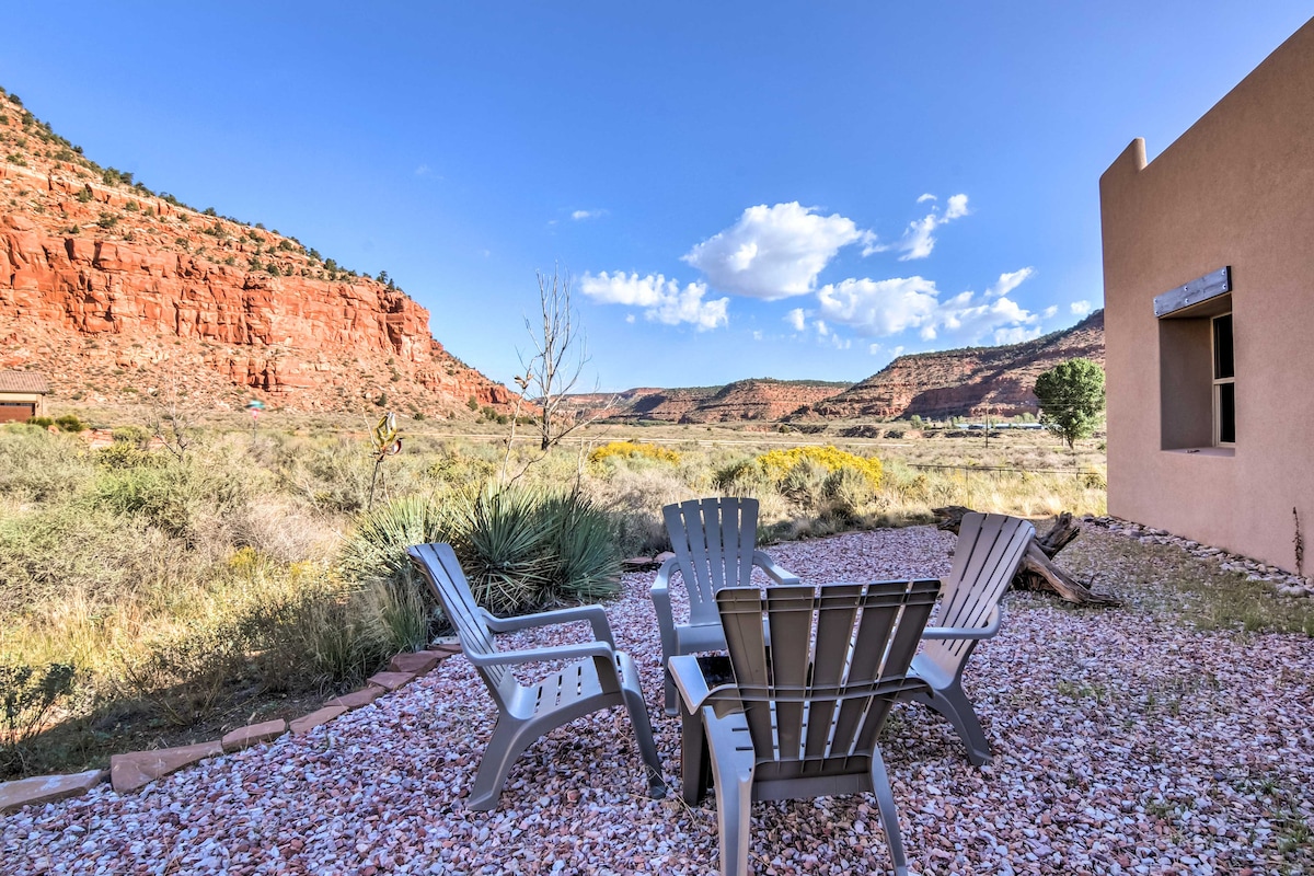 An outdoor seating area is set with four gray chairs arranged around a small gravel space. The background features expansive views of red rock formations and green vegetation under a clear blue sky, creating a serene and natural environment.