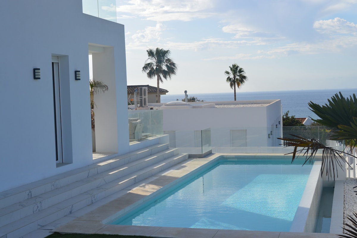 A modern villa is showcased with a private pool surrounded by sunlit terraces. Palm trees frame the background, with views extending toward the sea. Clear blue skies complement the contemporary architecture, enhancing the inviting outdoor space.