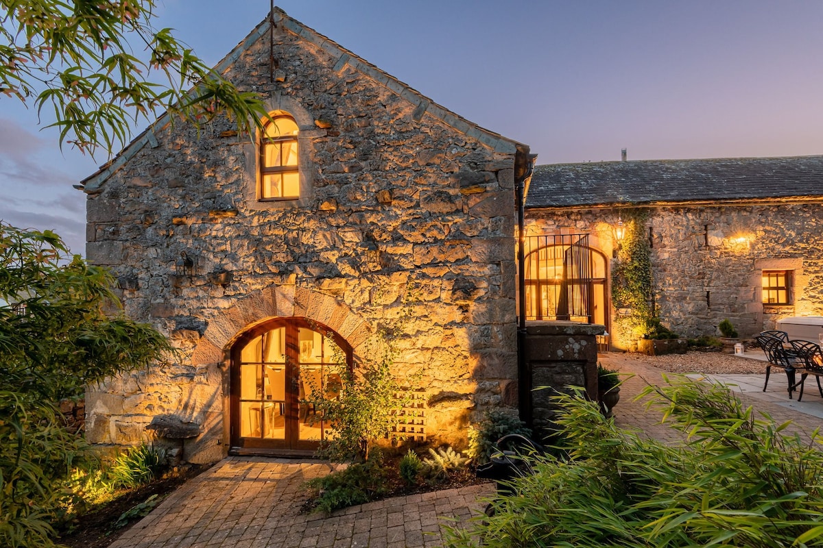The exterior of the barn conversion is illuminated by warm lights, highlighting the stone facade and arched doorway. Surrounding greenery adds a touch of nature, while the inviting entryway is framed by large windows, creating a welcoming atmosphere as dusk approaches.