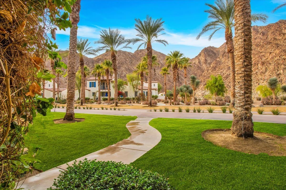 A winding pathway leads through a lush green lawn, framed by tall palm trees. In the background, rugged mountain peaks rise under a clear blue sky, complementing the serene landscape of the Legacy Villas community.