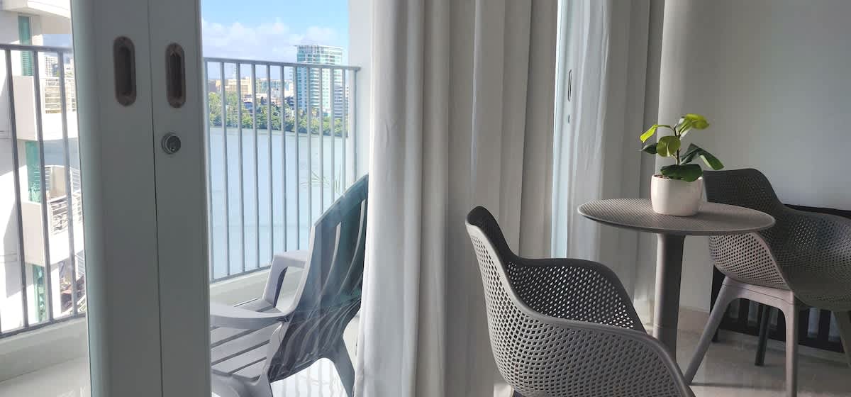 A private balcony is visible through glass doors, featuring a round dining table and two textured chairs. A potted plant adds a touch of greenery. Soft curtains frame the space, while the calm lagoon view is seen beyond the railing, enhancing the serene atmosphere.
