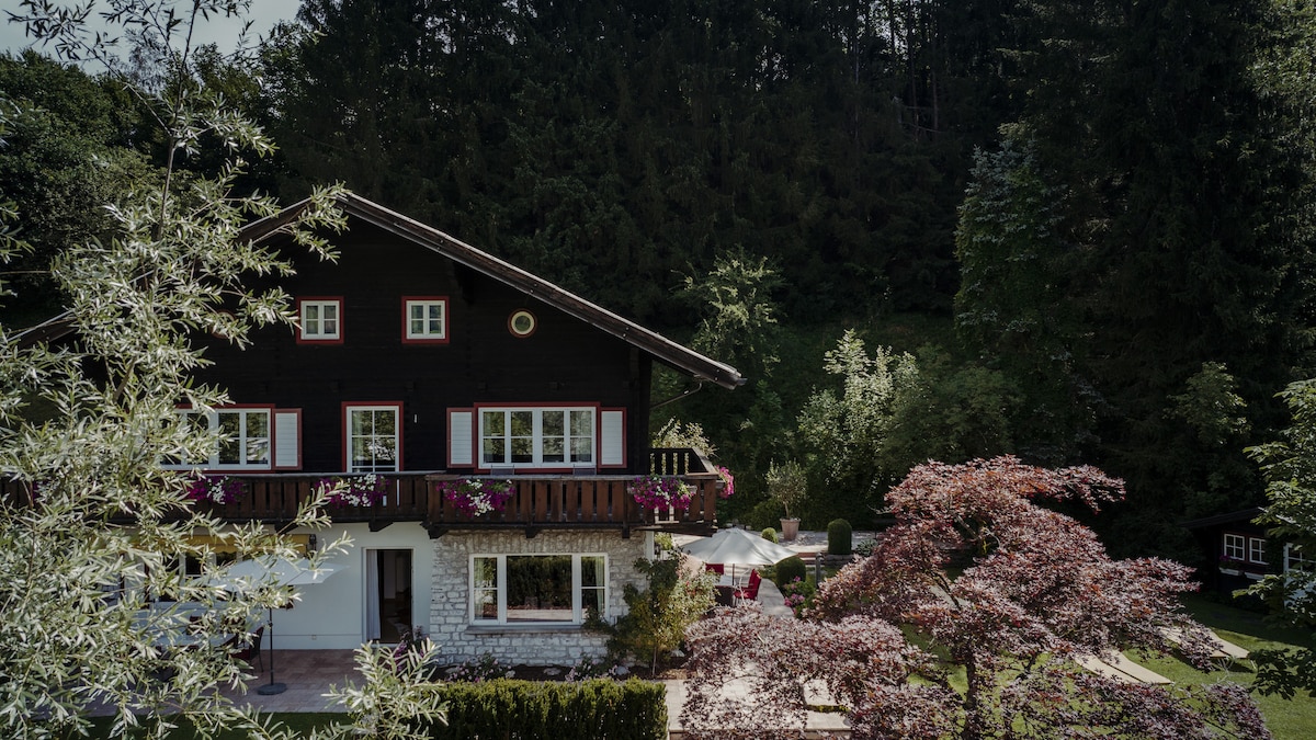 The two-story chalet is surrounded by lush greenery, featuring a blend of wooden and stone architecture. Colorful flowers adorn the balconies, while a patio area with an umbrella invites outdoor relaxation. A serene garden setting enhances the picturesque environment.