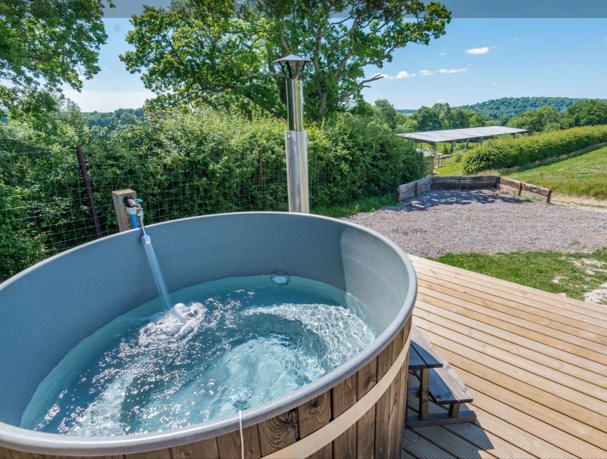 A wood-burning hot tub is placed on a deck, with clear water visible inside. Surrounding greenery frames the scene, while a pathway leads to open fields in the background, adding to the secluded atmosphere of the retreat.
