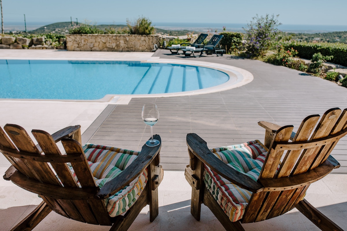 Two rustic wooden chairs with colorful cushions are positioned by a clean swimming pool. A crystal glass of wine rests on one chair, while the pool's blue water reflects sunlight. Lush greenery and distant hills are visible in the background.