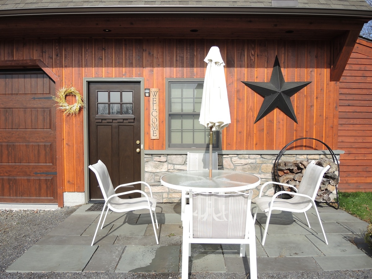 A charming entrance features a wooden door adorned with a welcoming sign. A round table with white chairs is set on a flagstone patio, complemented by a large umbrella. A rustic star decorates the wall, enhancing the inviting ambiance.