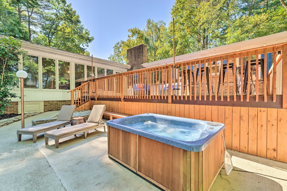 An outdoor area features a hot tub surrounded by a wooden deck. Two lounge chairs are positioned nearby, offering a place to relax. The backdrop consists of greenery and a large sunroom with expansive windows, creating a connection to nature.