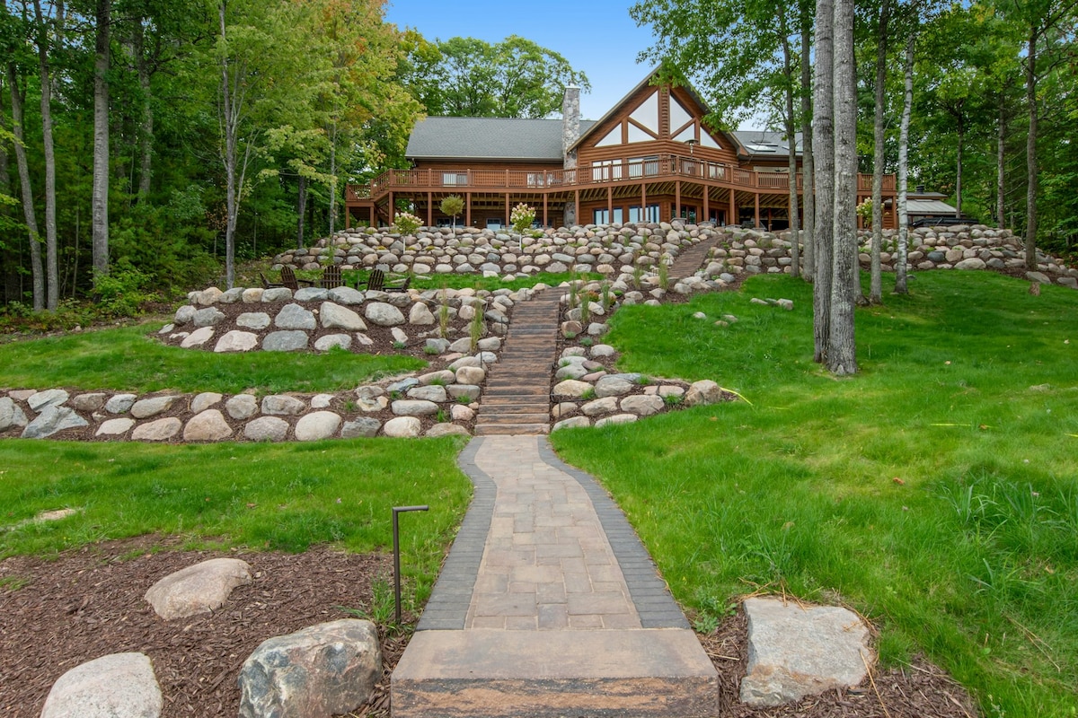 The exterior of the log chalet is surrounded by lush greenery and features a stone pathway leading to a spacious deck. Natural stone landscaping integrates with the terrain, and the environment showcases a serene, wooded backdrop.