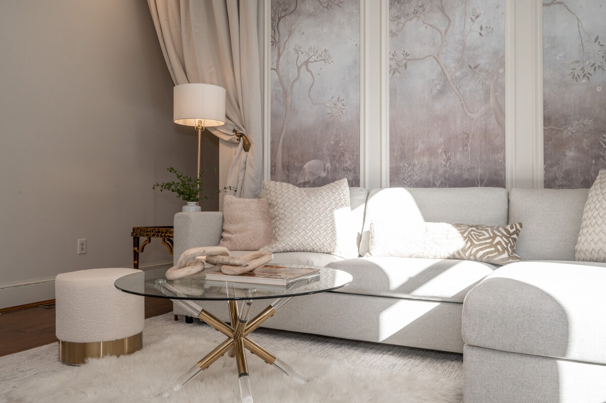 A modern seating area is presented with a light-colored sectional sofa adorned with decorative pillows. A round glass coffee table rests on a soft rug, complemented by a stylish lamp and natural greenery, while textured artwork adds visual interest to the wall.