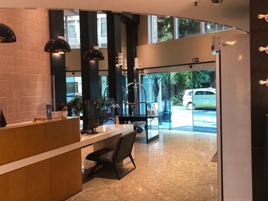 The lobby area is designed with modern finishes, featuring a reception desk made of wood and a sleek black chair. Large glass doors open to the street, allowing natural light to brighten the space. Potted plants add a touch of greenery.