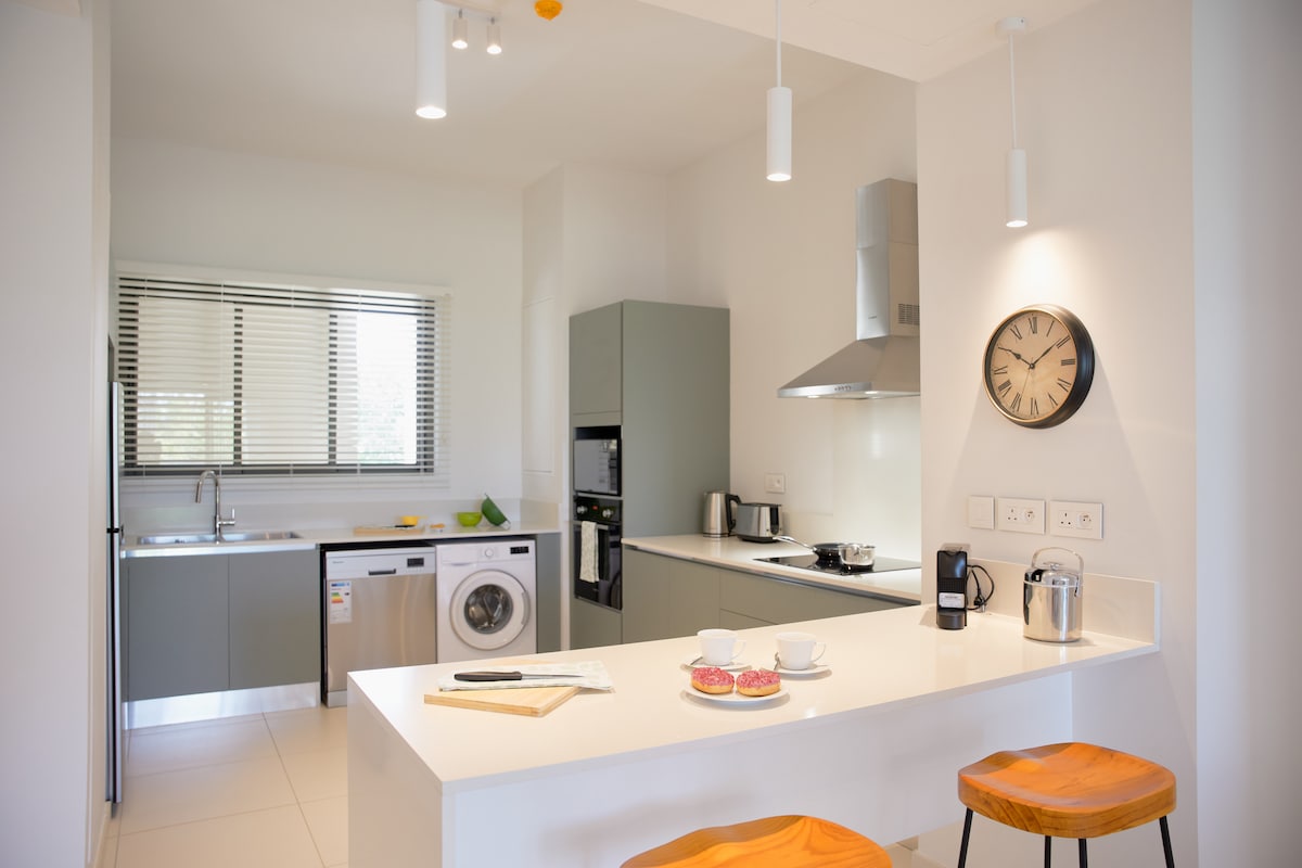 An open-plan kitchen is equipped with modern appliances, including a refrigerator, washing machine, and coffee maker. A kitchen island with two wooden stools provides a casual dining option. Natural light fills the space through large windows, enhancing the clean, minimalist design.