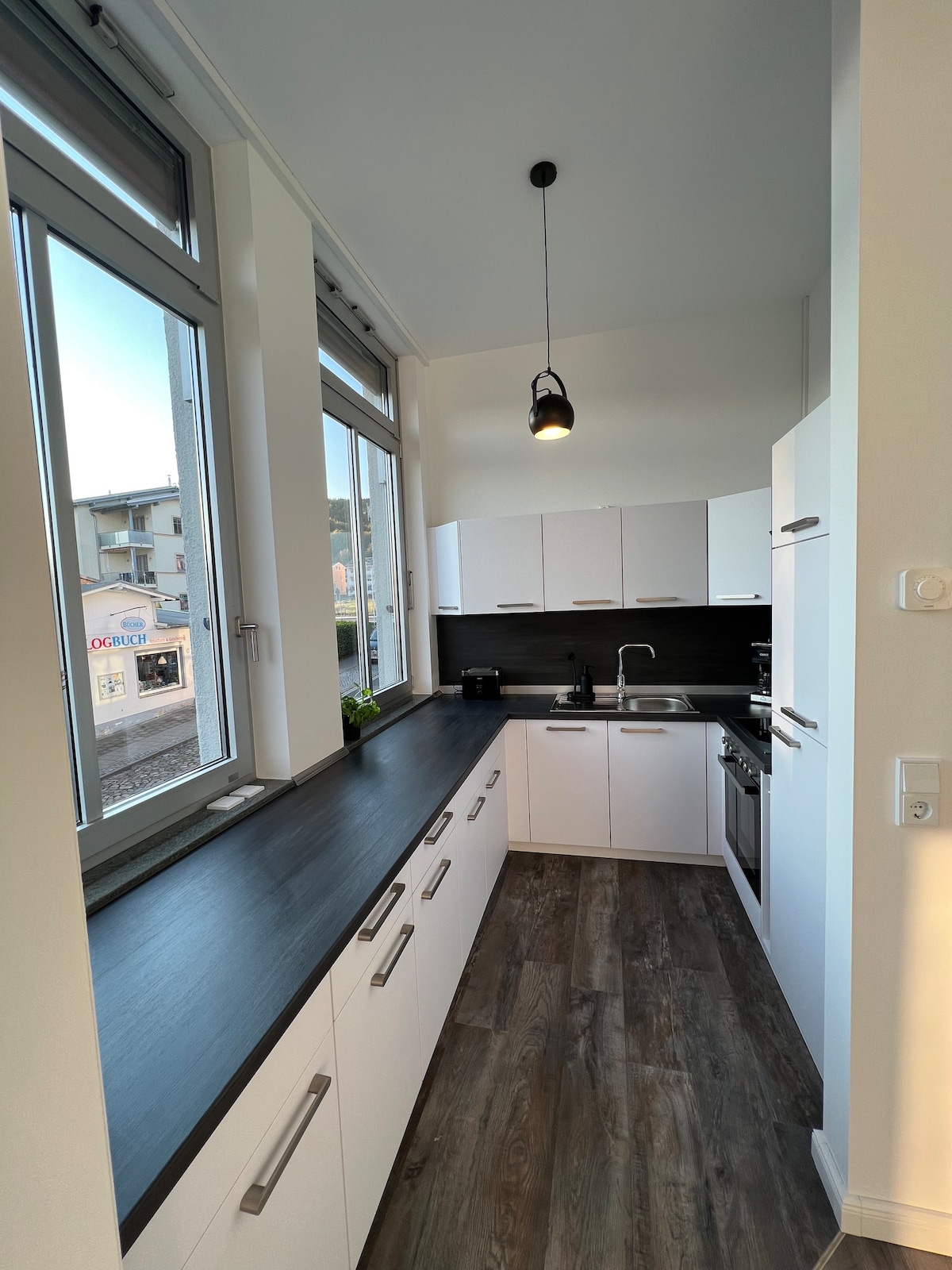 The open kitchen is designed with modern white cabinetry and a dark countertop. Large windows allow natural light to fill the space, illuminating the cooking area, which contains a hob, oven, and sink. Selected kitchen utensils are neatly arranged, providing a functional cooking environment.
