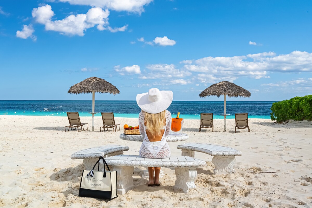 A person is seated at a round white stone table on a sandy beach, gazing out at the ocean. Two thatched umbrellas provide shade for loungers placed nearby, with a beach bag and refreshments arranged on the table.