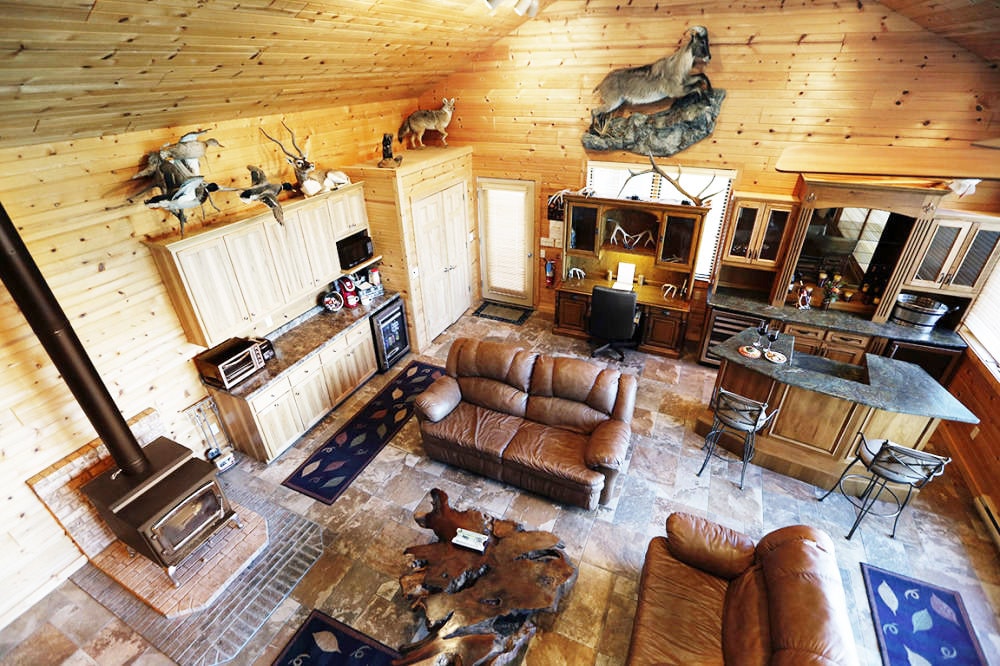 An open living space features a rustic wooden interior with a stone floor. A plush brown leather seating arrangement faces a modern stove, while a kitchen area with wooden cabinetry and bar seating is visible in the background. Taxidermy wildlife accents the walls.