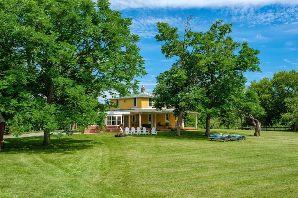 A yellow farmhouse is surrounded by lush green grass and large trees, creating a serene outdoor environment. White lawn chairs are arranged in groups on the patio, while additional seating is visible nearby. The clear blue sky adds to the tranquil atmosphere.