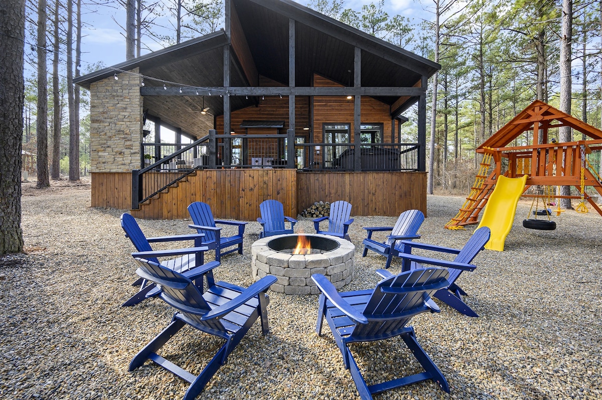 An outdoor seating area is arranged around a stone firepit, with blue Adirondack chairs set in a circle. The cozy cabin, featuring large windows and a sloped roof, is framed by tall pine trees. A playset with a slide and swings is visible nearby.