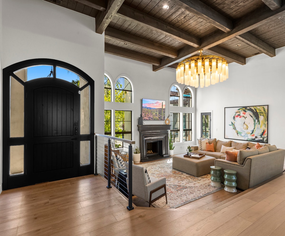 A spacious living area features tall arched windows that allow natural light to fill the room. A plush sectional couch is positioned around a coffee table, complemented by decorative pillows. A modern chandelier hangs from the ceiling above, and a flat-screen TV is mounted above a stylish fireplace.