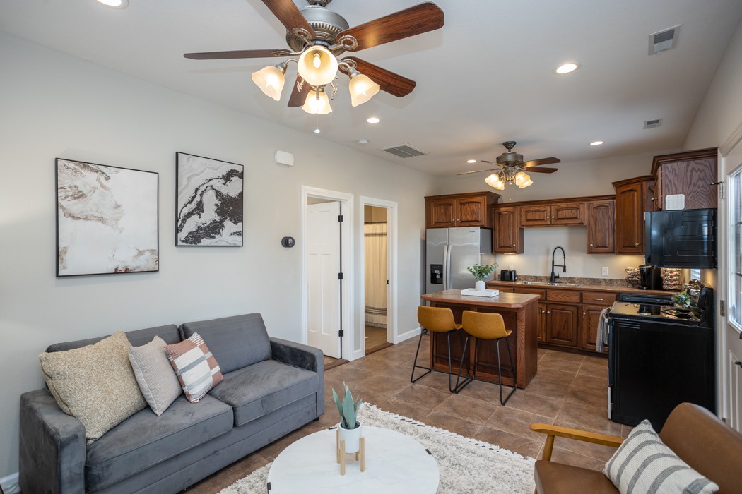 An open-concept living area features a cozy couch with decorative pillows, complemented by a round coffee table. A well-equipped kitchen showcases wooden cabinetry, stainless steel appliances, and an island with bar stools, enhancing the functional design of the space.