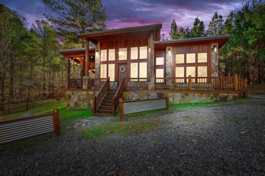 The cabin features a warm wooden façade with large windows, allowing for natural light to illuminate the interior. A stone path leads to the entrance, framed by lush greenery and a gravel driveway, creating a serene and inviting atmosphere among the surrounding trees.