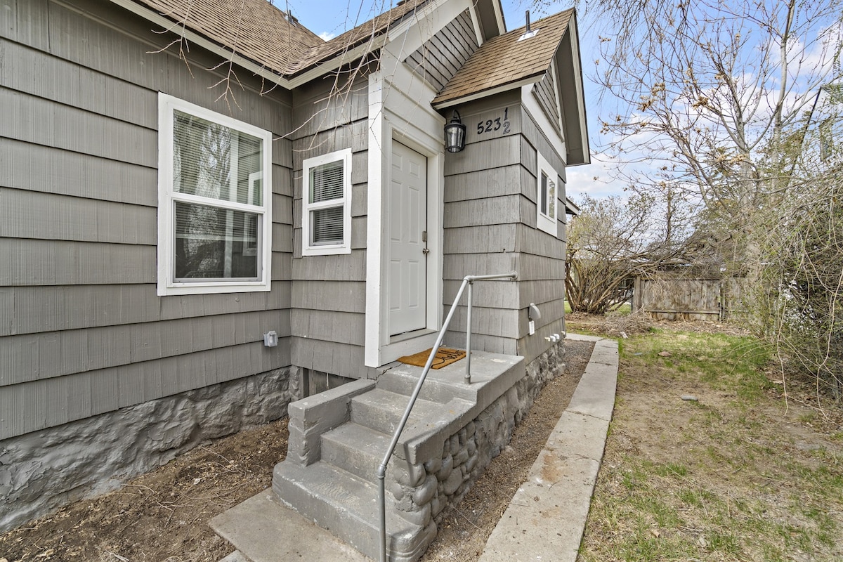 The exterior entrance features a concrete pathway leading to a white door, bordered by gray siding and a shingled roof. A lantern-style light fixture is mounted beside the door, and a doormat is placed at the entryway. Surrounding vegetation adds a natural element.