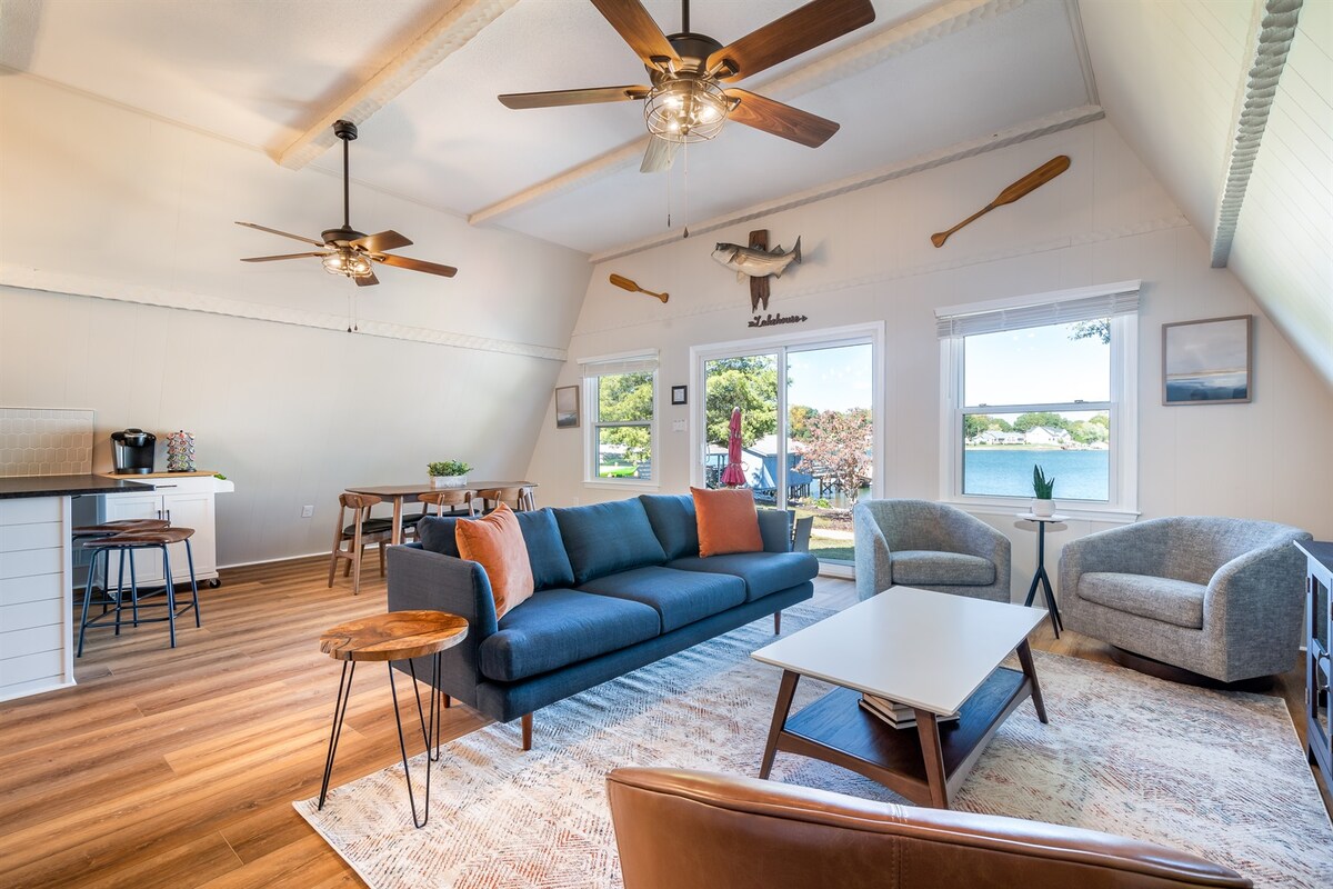 An inviting family room is displayed with modern furnishings, including a spacious blue sofa and two comfortable armchairs. Large windows provide views of Lake Norman. A dining table is visible in the background, along with a ceiling fan and decorative wall art, enhancing the space's relaxed ambiance.