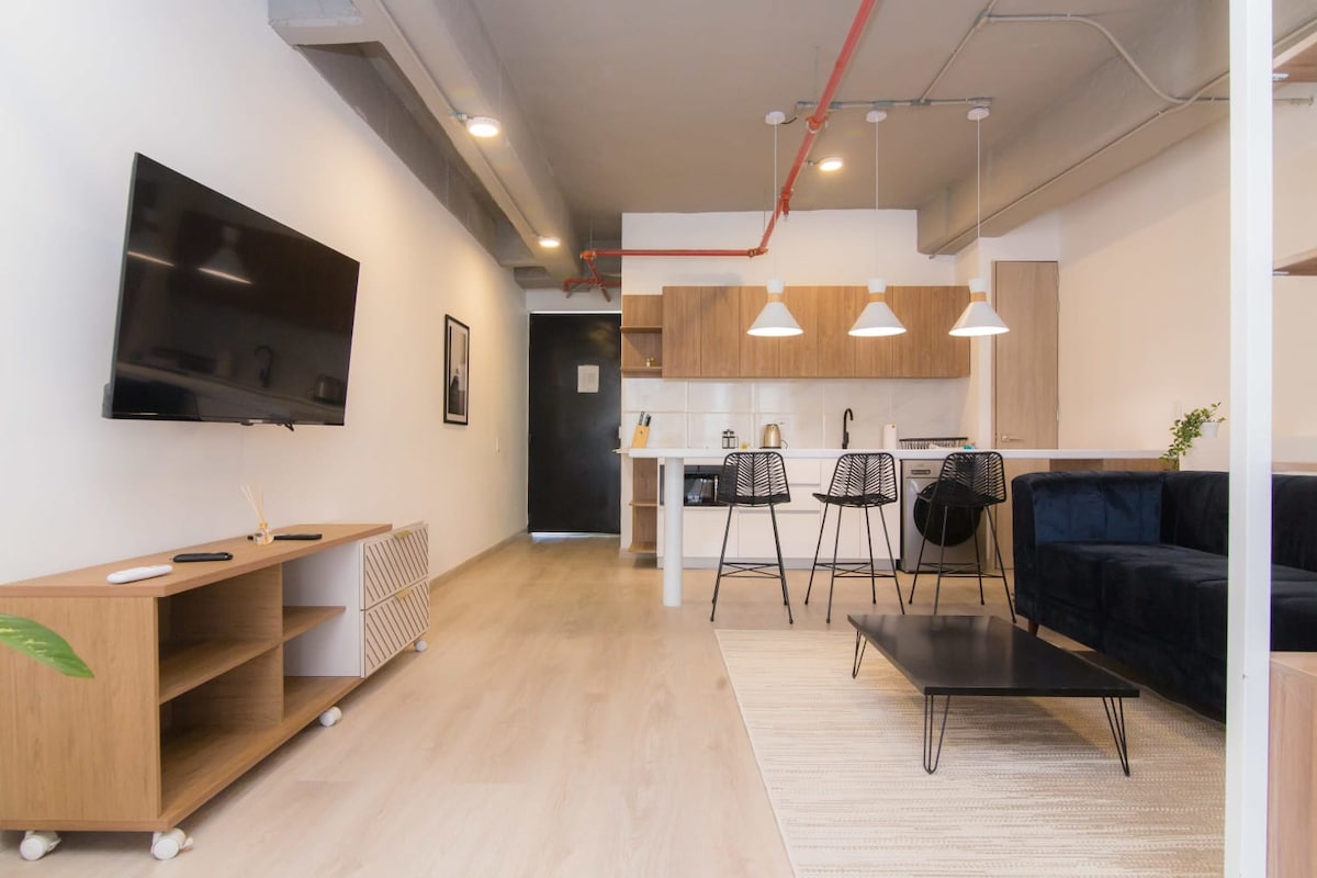 A spacious living area features light wooden flooring, with a modern kitchen visible in the background. Black dining chairs surround a sleek table, while a dark sofa adds depth to the open layout. A flat-screen TV is mounted on the wall, enhancing the functional space.