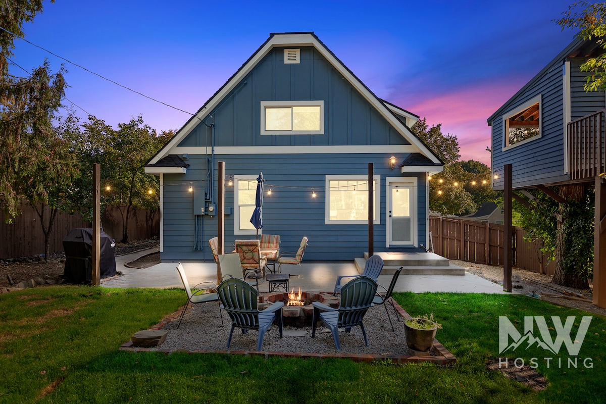 The backyard features a firepit surrounded by several chairs, providing a cozy gathering spot. The blue farmhouse exterior is visible, complemented by string lights above. A treehouse peeks from the side, enhancing the inviting atmosphere of the space.
