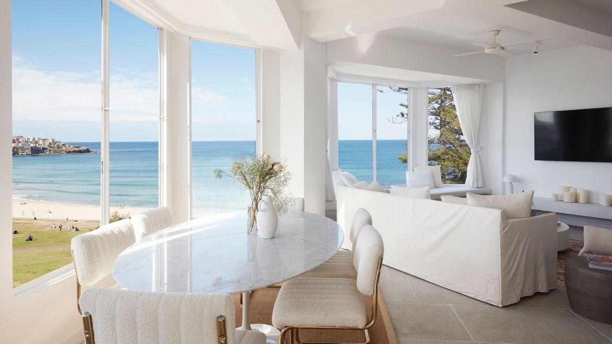 An open-plan living and dining area is showcased, featuring large windows that frame ocean views. A round marble dining table is surrounded by light-colored chairs, while a cozy seating area with white sofas invites relaxation. Sunlight floods the space, creating a bright atmosphere.