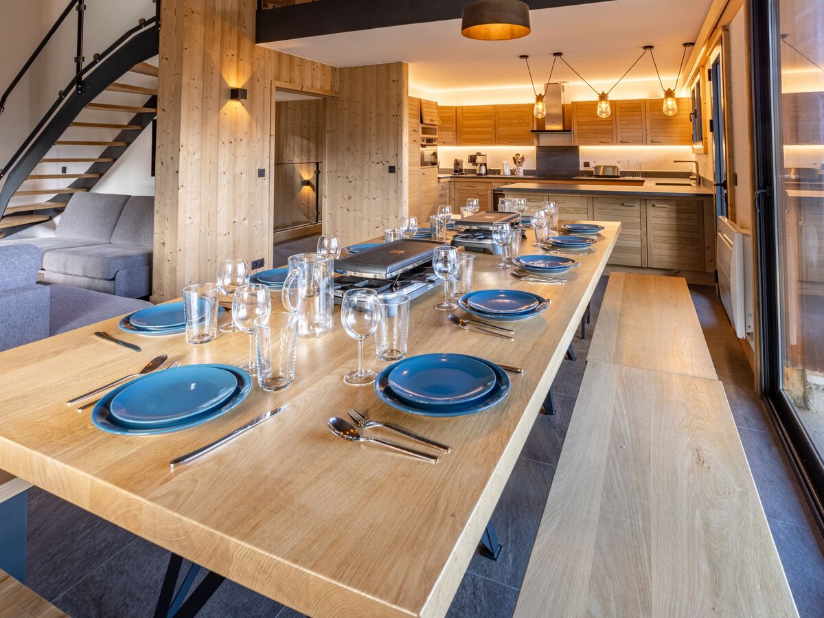 A spacious dining area is featured, with a long wooden table set for guests. Each place setting includes blue plates and glassware. Modern light fixtures illuminate the space, while a well-equipped open kitchen is visible in the background.