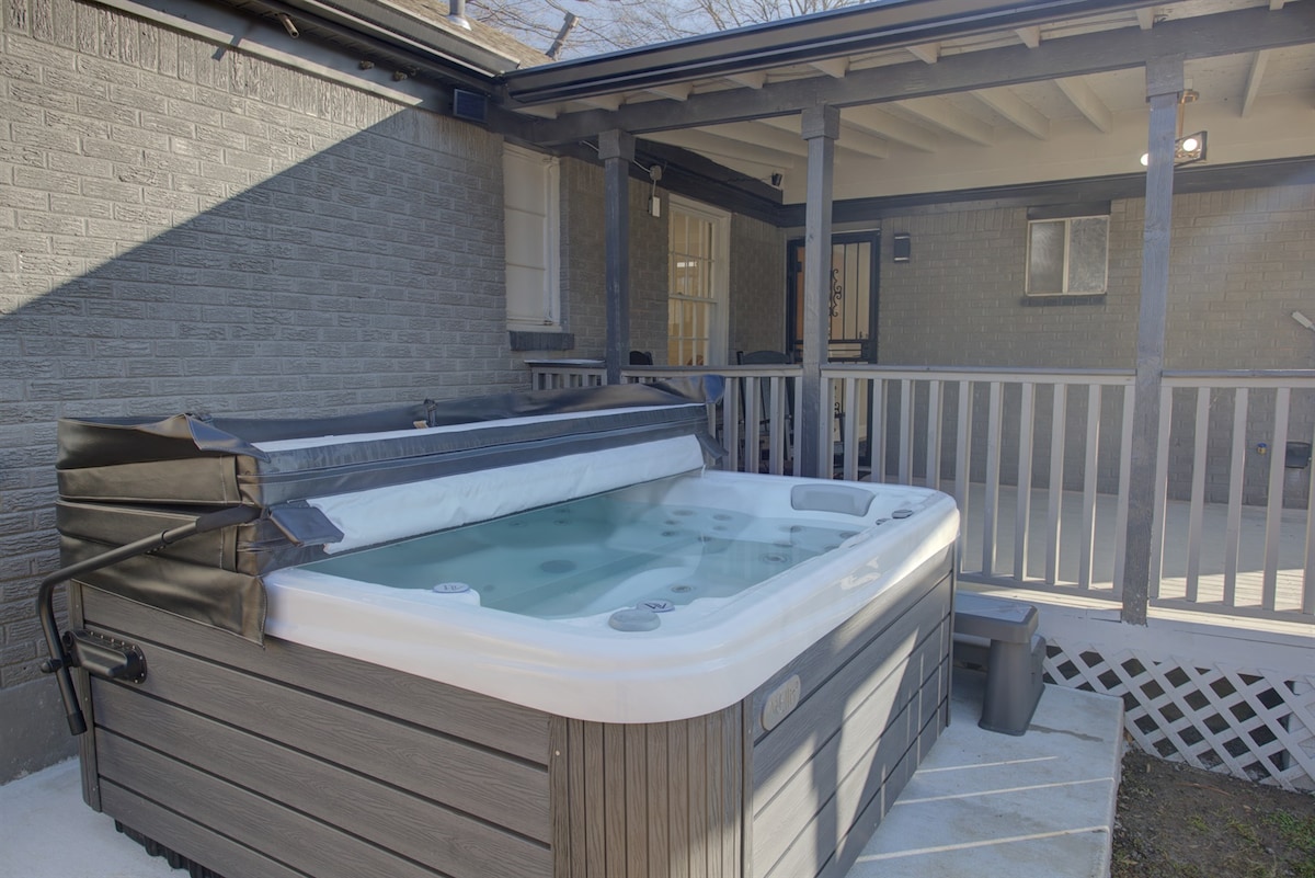 An outdoor hot tub is situated on a raised wooden deck, featuring a covered area and white railings. The tub is filled with clear water, and the cover is partially on, allowing for easy access. Sunlight illuminates the surrounding area.