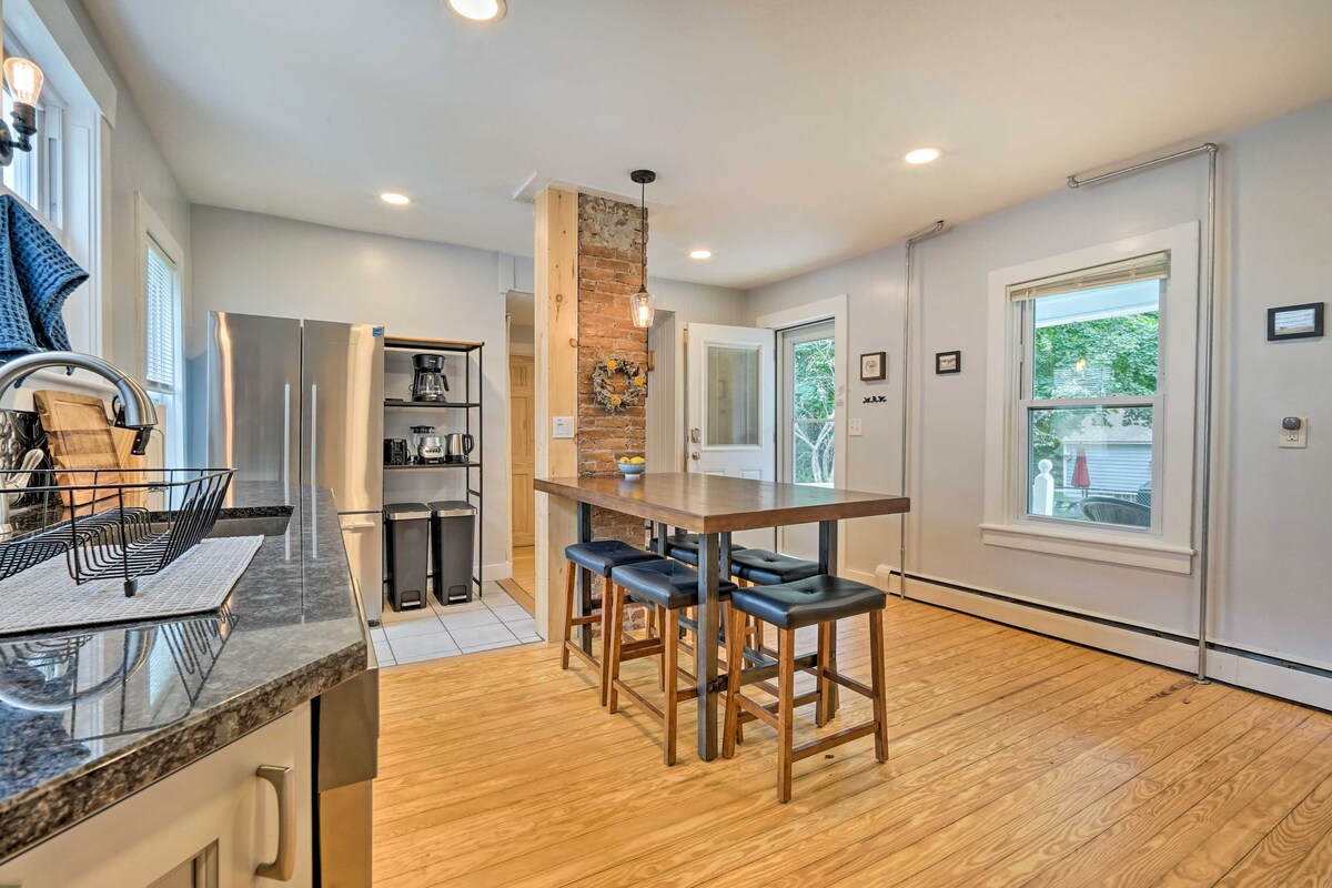 A bright kitchen area features modern appliances, including a refrigerator and coffee maker. A central dining table with stools provides a gathering space. Large windows allow natural light to illuminate the wooden floors and reveal views of the outdoor space.
