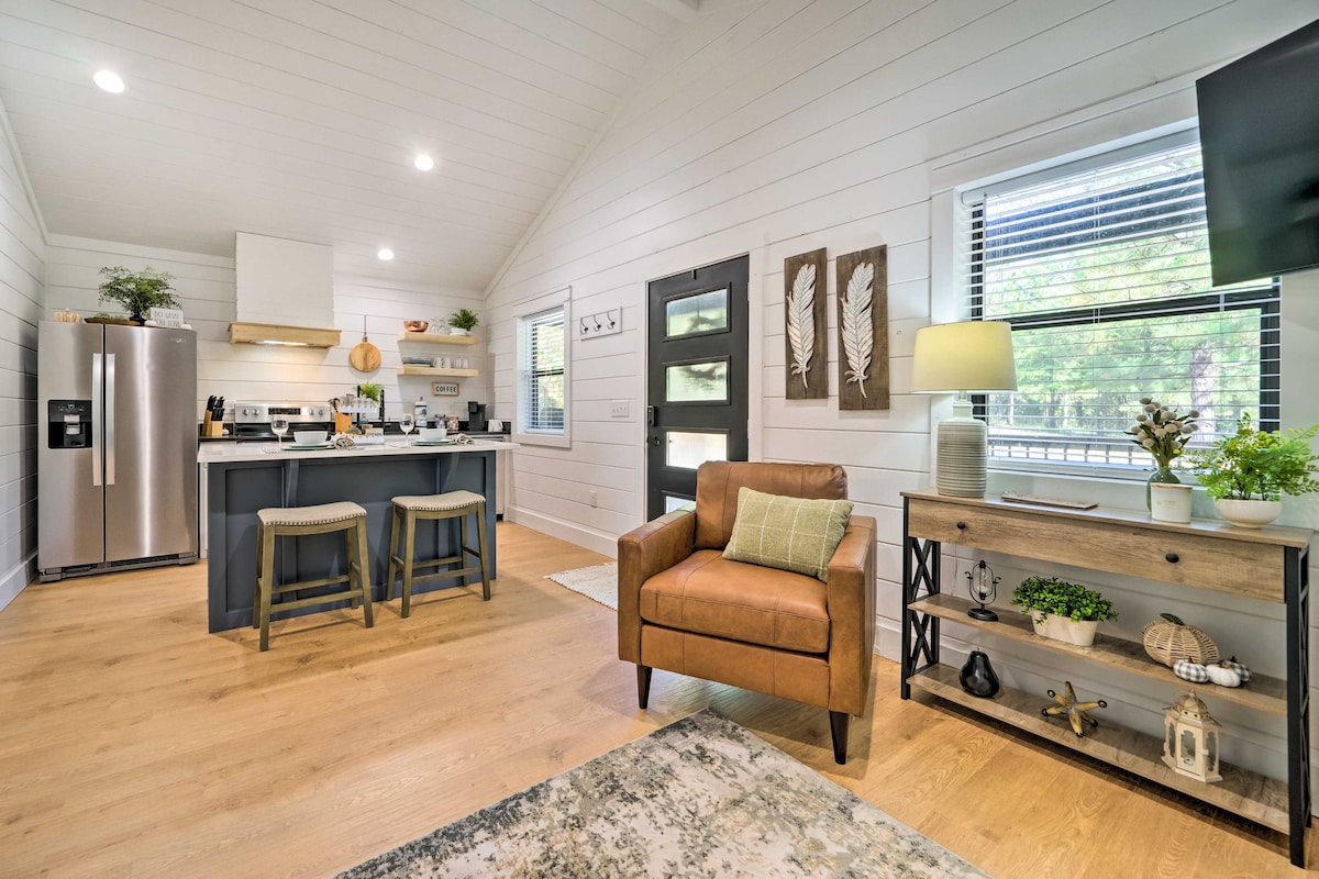 An open-concept living area is featured, showcasing a modern kitchen with stainless steel appliances and a breakfast bar. A comfortable brown armchair is positioned next to a decoratively arranged console table. Natural light fills the space through large windows, enhancing the airy atmosphere.