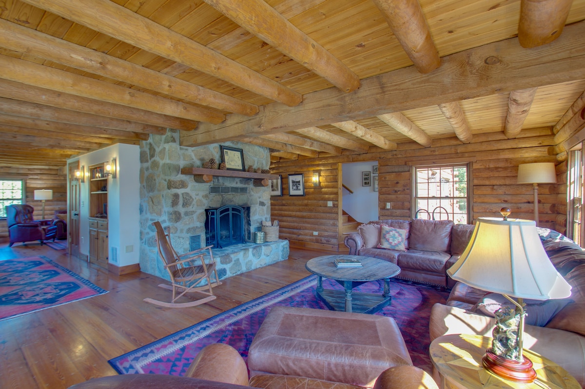 A spacious living area is highlighted by warm wooden beams and a stone fireplace. Comfortable seating arrangements surround a central coffee table, with a rug adding warmth. Natural light enters through large windows, illuminating the rustic decor throughout the room.