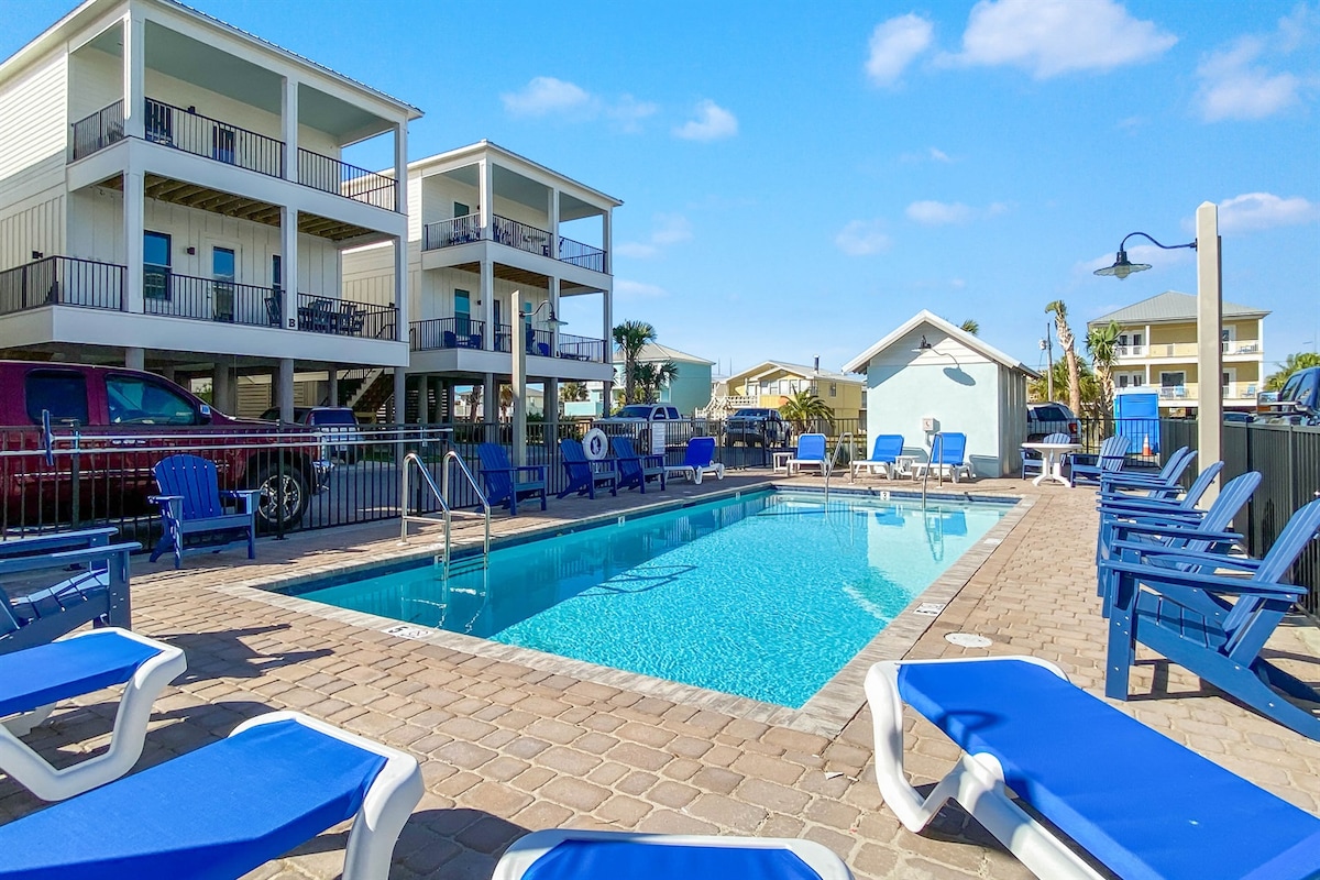 A communal pool area is surrounded by lounge chairs with blue cushions, allowing for relaxation under the sun. The pool is bordered by a stone deck, and nearby buildings display a coastal style, with two stories visible in the background along with palm trees.