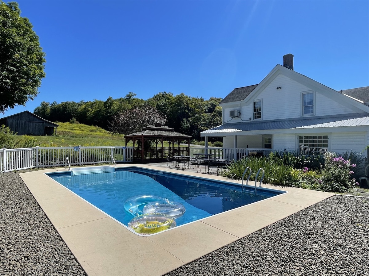 An in-ground pool is surrounded by a flagstone patio and flower gardens. A gazebo with outdoor seating is visible next to the pool, which features inflatable floats resting on the water. The white house with a metal roof stands in the background, complemented by clear blue skies.