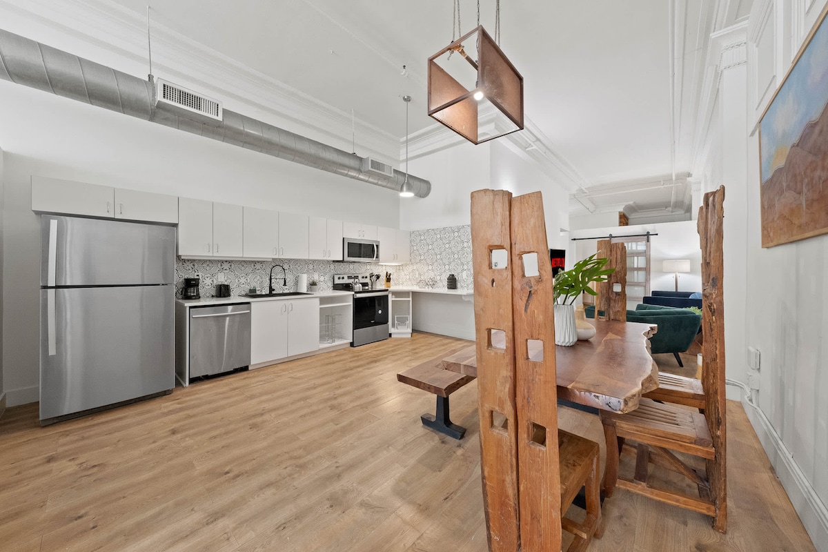 A spacious kitchen area features modern stainless steel appliances, including a refrigerator and oven. A large wooden dining table with unique chairs sits in the foreground, accompanied by a potted plant. The open floor plan enhances connectivity between the kitchen and dining space.