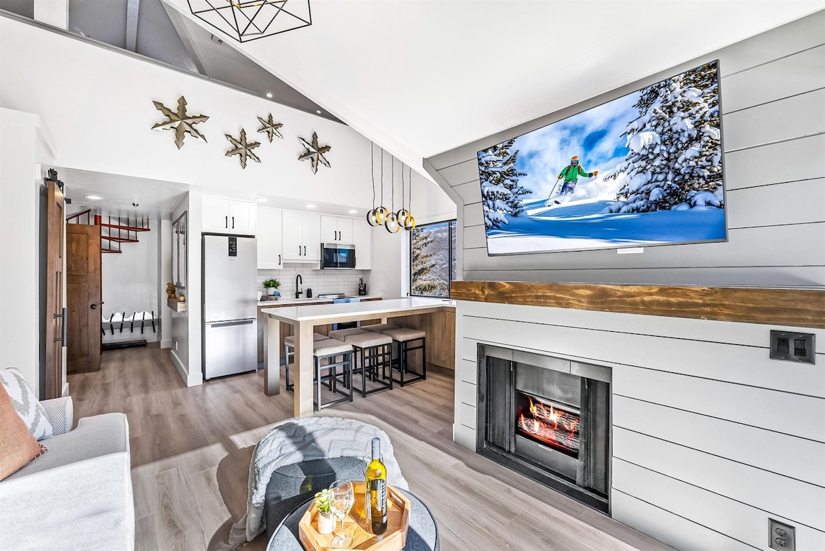 A spacious living area features modern decor with vaulted ceilings and large windows showcasing views of the mountains. A stylish fireplace is complemented by a mounted Smart TV, and a dining island with seating overlooks a well-equipped kitchen. Decorative stars add a unique touch.
