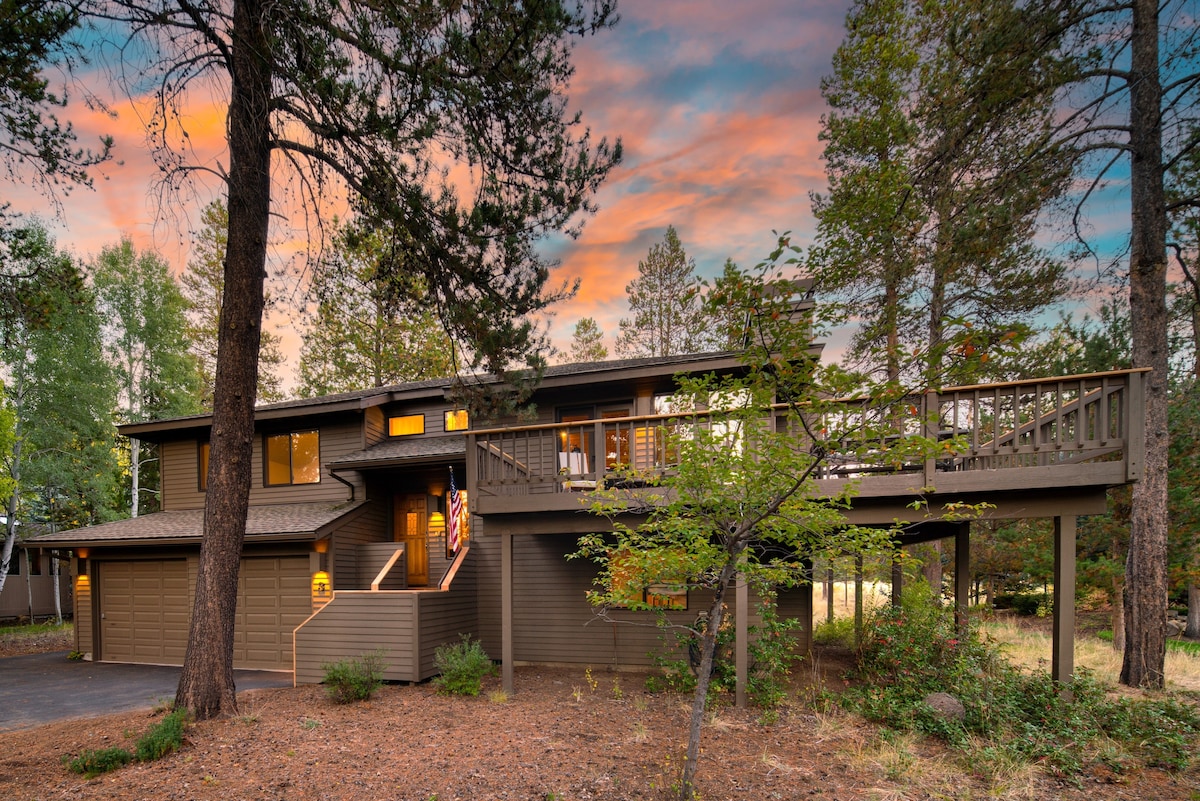 A modern mountain home is surrounded by trees, featuring a spacious deck that extends from the upper level. The unique architecture is complemented by evening colors in the sky, creating a serene backdrop for the entrance, which is warmly lit.