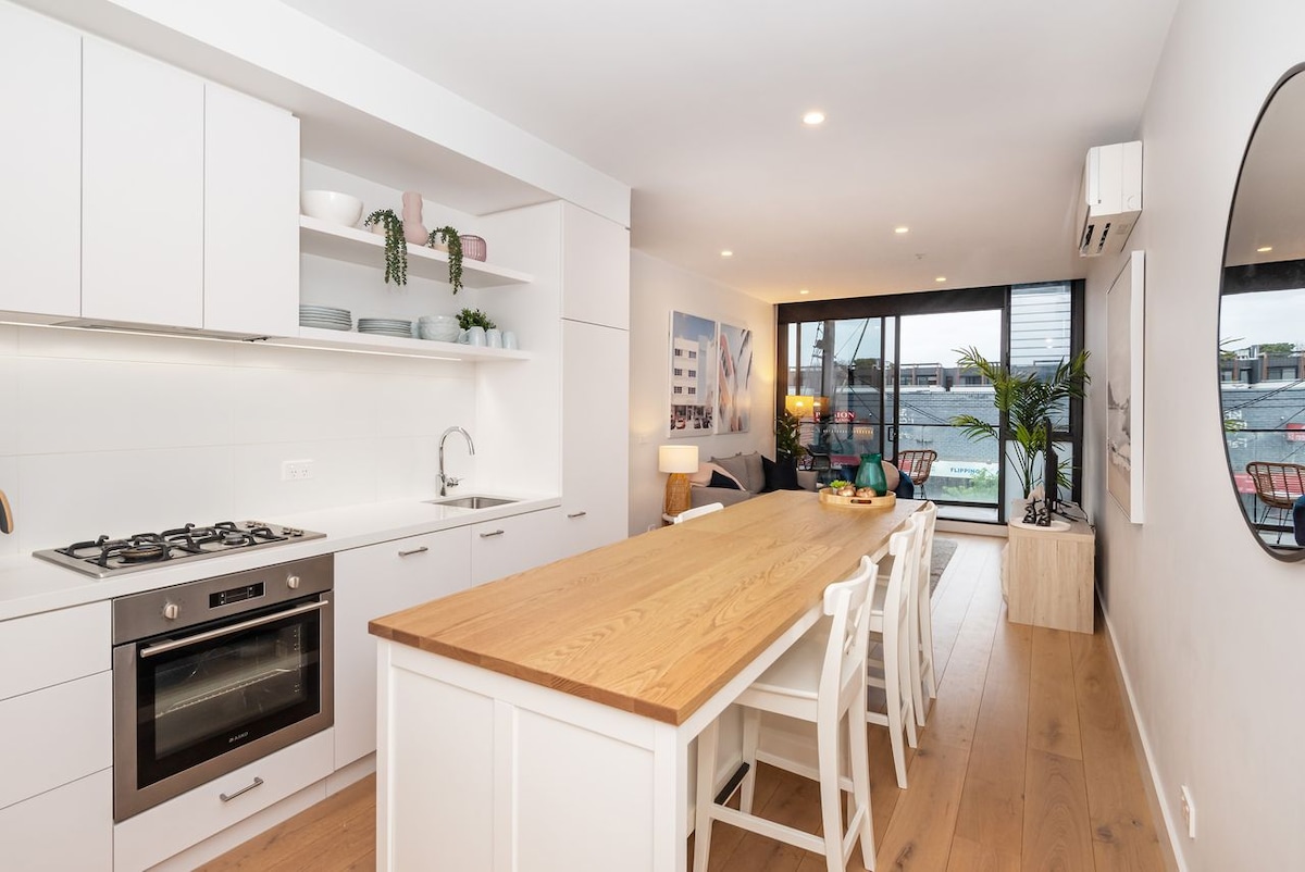 A modern kitchen and dining area features a long wooden table surrounded by white chairs. Sleek cabinetry and open shelving display various dishware. Natural light enters through large glass doors, with comfortable seating visible in the background, creating a welcoming and functional space.